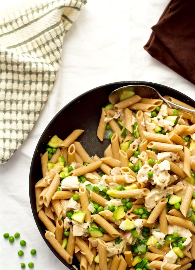 A spoon resting in a skillet of pasta with chicken, zucchini and peas.