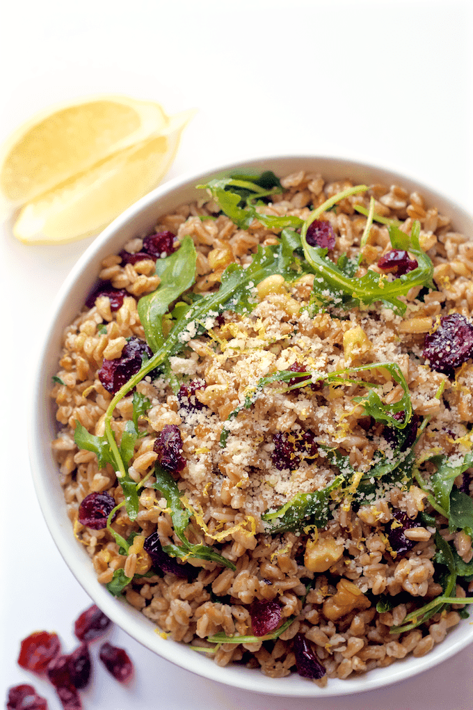 A farro salad with arugula, walnuts and dried cranberries in a large white bowl with lemon zest and Parmesan cheese sprinkled on top.