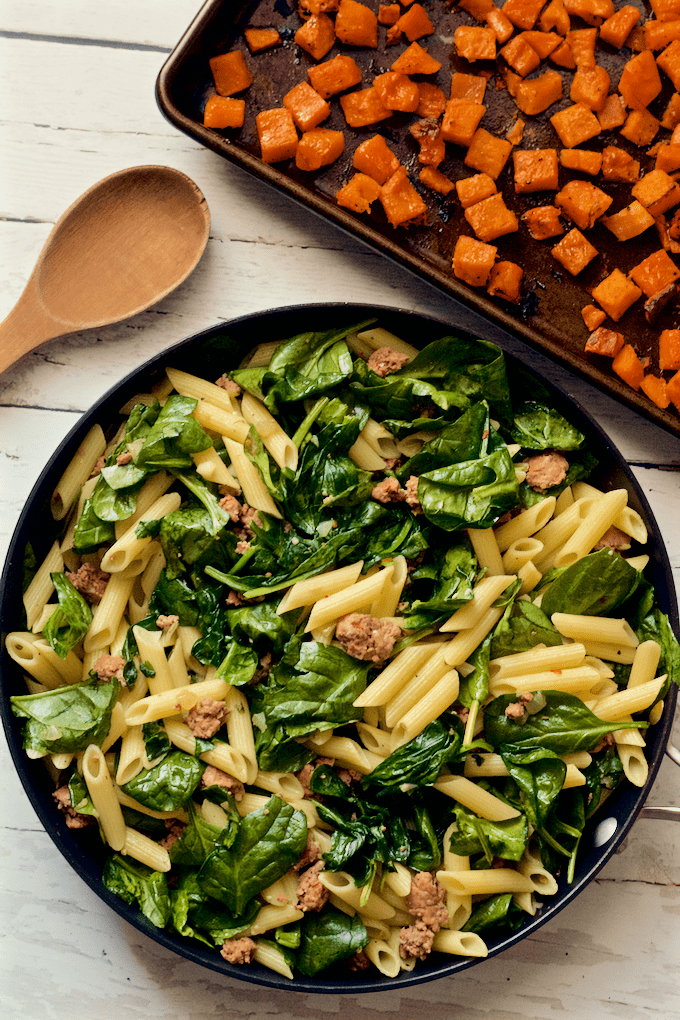 A sheet pan of roasted butternut squash alongside a skillet with a mixture of pasta, spinach and turkey sausage.