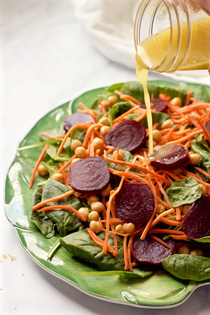 A lemon vinaigrette being poured from a glass jar onto a salad of spinach, chickpeas, beets and matchstick carrots.