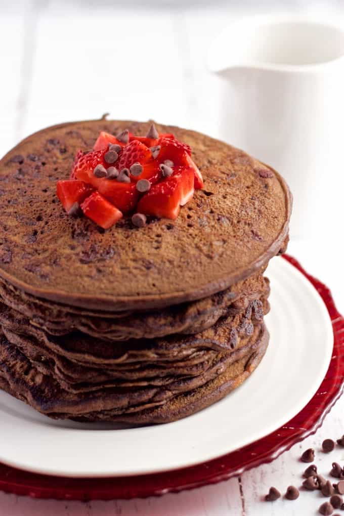 These healthy chocolate pancakes are whole wheat, naturally sweetened and made without butter or oil. They're great for a fun and special breakfast! #chocolatebreakfast #chocolatepancakes #pancakes A stack of chocolate pancakes on a white plate with diced strawberries and mini chocolate chips on top.
