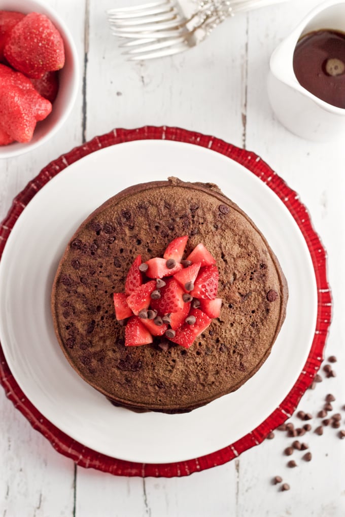 Overhead of a stack of double chocolate pancakes on a white plate with diced strawberries and mini chocolate chips on top.