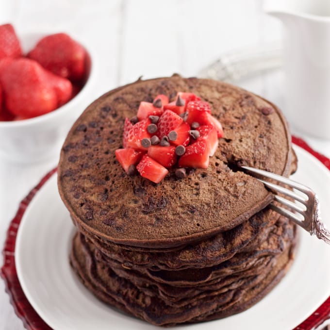 A fork cutting into a stack of chocolate pancakes on a white plate with diced strawberries and mini chocolate chips on top.
