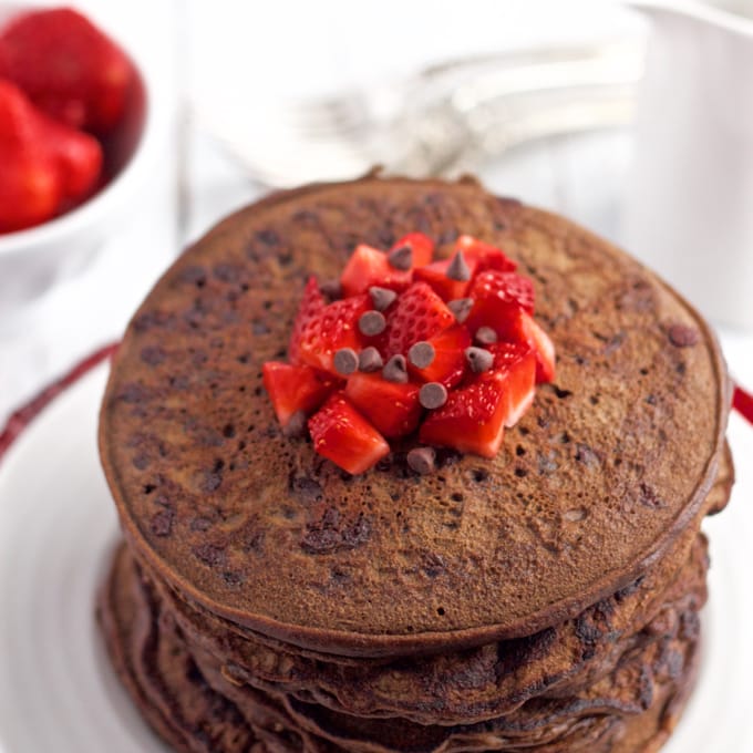 A big stack of double chocolate pancakes on a white plate with diced strawberries and mini chocolate chips on top and strawberries and syrup in the background.