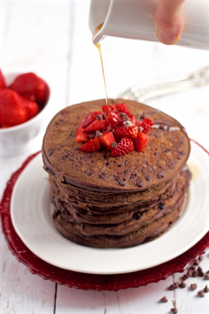 Maple syrup being drizzled over a stack of chocolate pancakes on a white plate with diced strawberries and mini chocolate chips on top.