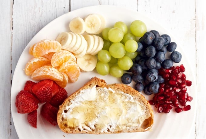 A piece of toasted bread with cream cheese and honey on it on a plate with a colorful array of fresh fruits.