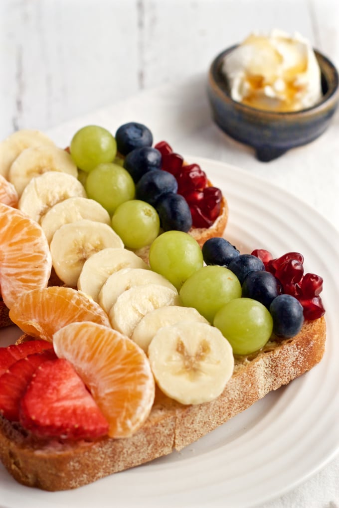 Close up of two pieces of toast on a plate with cream cheese topped with rows of rainbow colored fruit.