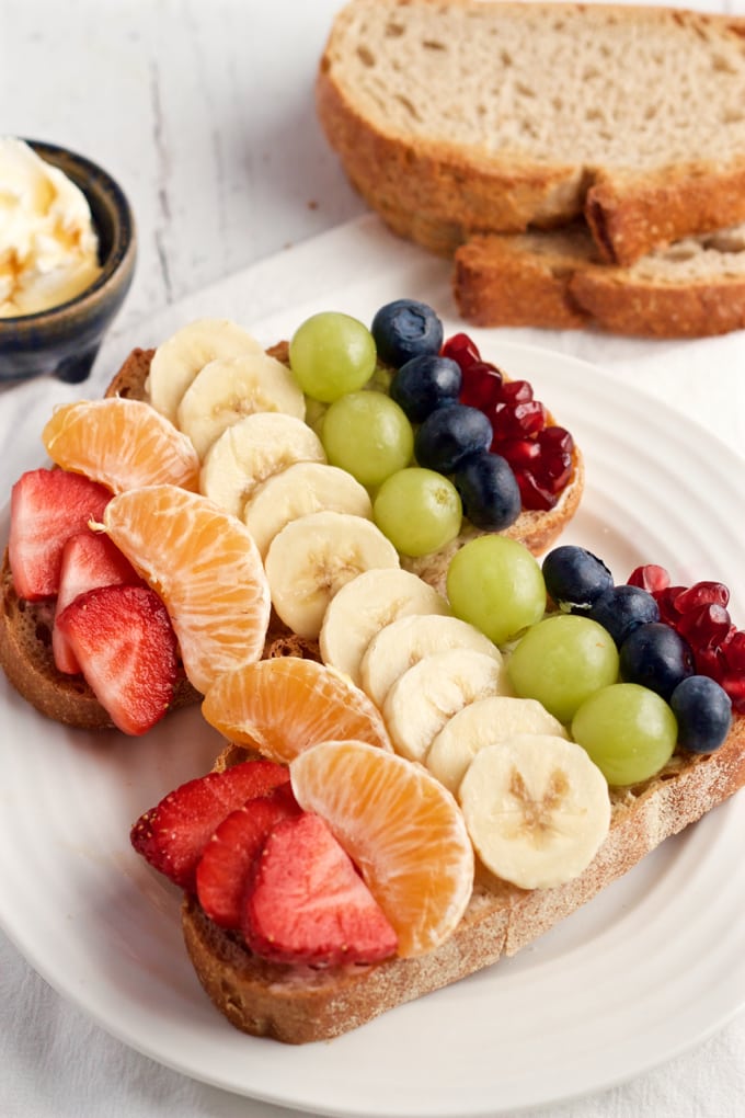Two pieces of toast on a plate with cream cheese topped with rows of rainbow colored fruit.