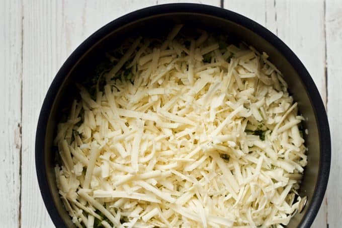 Shredded white cheddar cheese on top of chopped broccoli added to a pot of steamed brown rice before being mixed in.