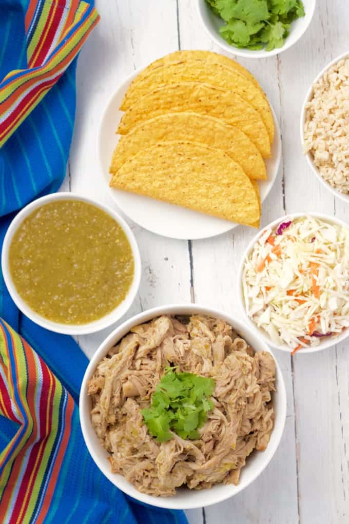 Slow cooker salsa verde shredded pork is great for tacos, rice bowls, quesadilla and nachos! #slowcooker #crockpot #porkrecipes #porktacos A taco bar set out with a plate of tacos, a bowl of steamed rice, cabbage, salsa verde shredded pork and a bowl of salsa verde.