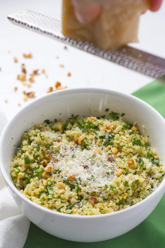 Fresh Parmesan cheese being grated over a bowl of quinoa spinach salad with pesto and nuts.