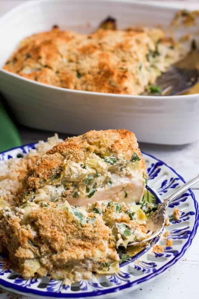 A baked spinach artichoke chicken breast served with rice on a blue and white plate and a casserole dish in the background