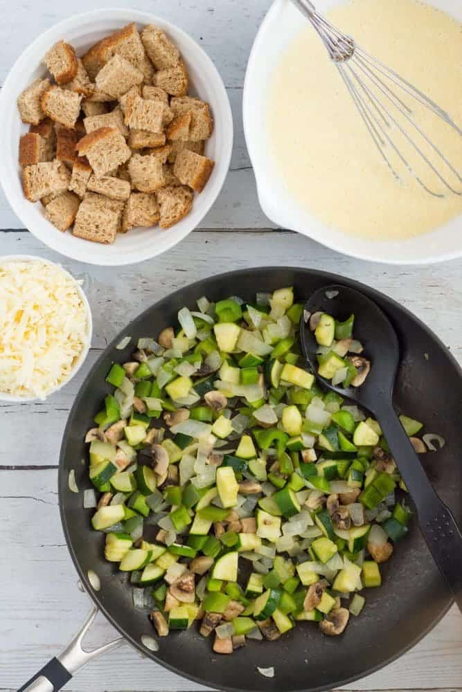 Veggie frittata ingredients prepped and laid out on a counter, including a skillet of tender veggies, cubed bread chunks, shredded cheese and a bowl of whisked eggs