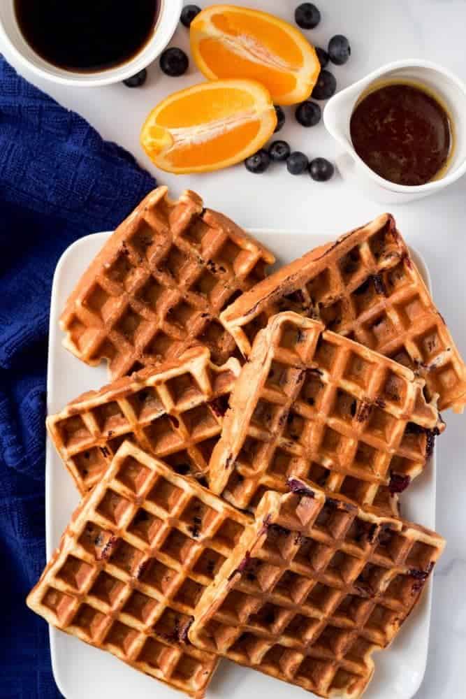 A platter of waffles being served with maple syrup, blueberries, oranges and coffee nearby