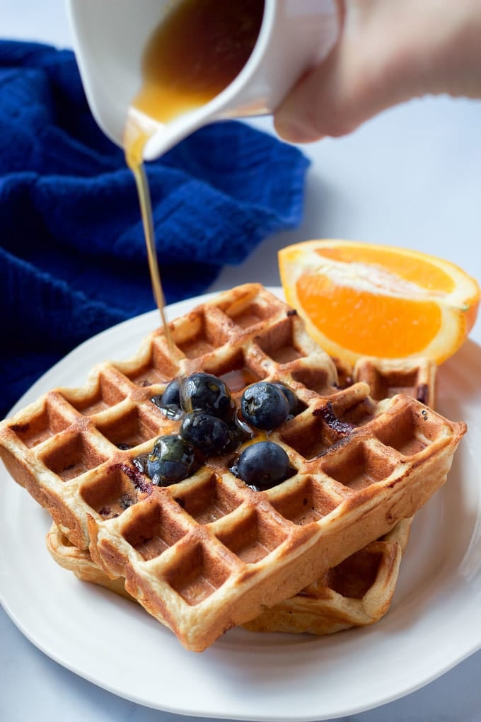 Maple syrup being drizzled over whole wheat waffles with blueberries on top