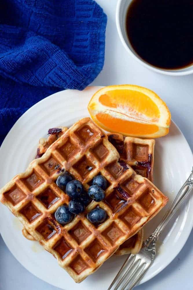 A stack of two blueberry waffles served on a plate with orange slices and fresh blueberries