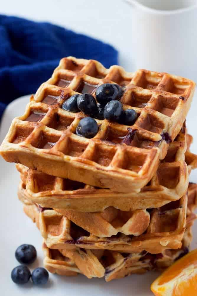 A tall stack of blueberry waffles with extra blueberries on top and nearby