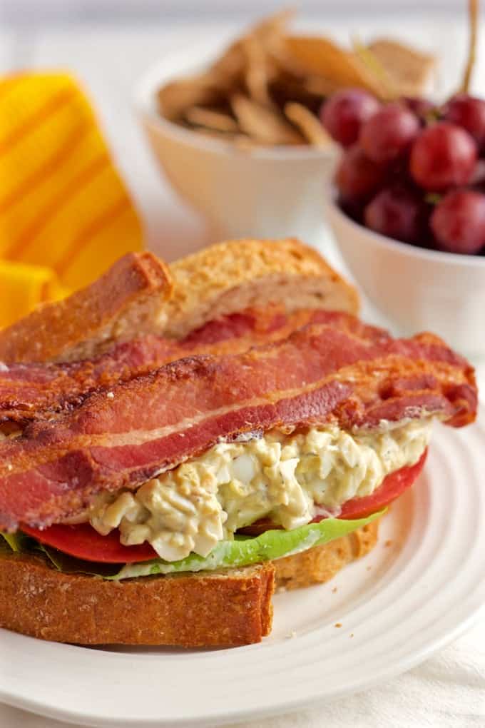 A creamy egg salad BLT sandwich with fresh tomatoes, soft Bibb lettuce and crispy strips of bacon makes for a stellar lunch sandwich! A bacon, lettuce and tomato egg salad sandwich on bakery bread with red grapes and chips in bowls in the background.