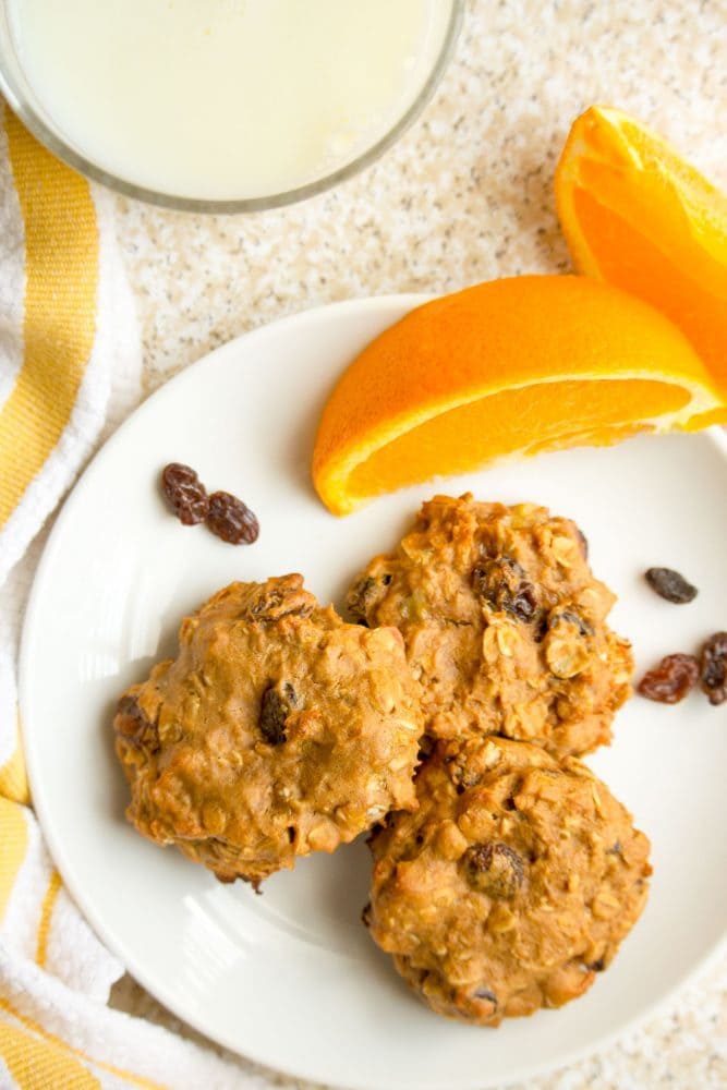 A plate full of breakfast cookies with orange slices, raisins and a glass of milk nearby
