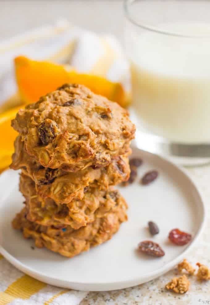 A stack of oatmeal raisin cookies on a white plate with milk and oranges in the background