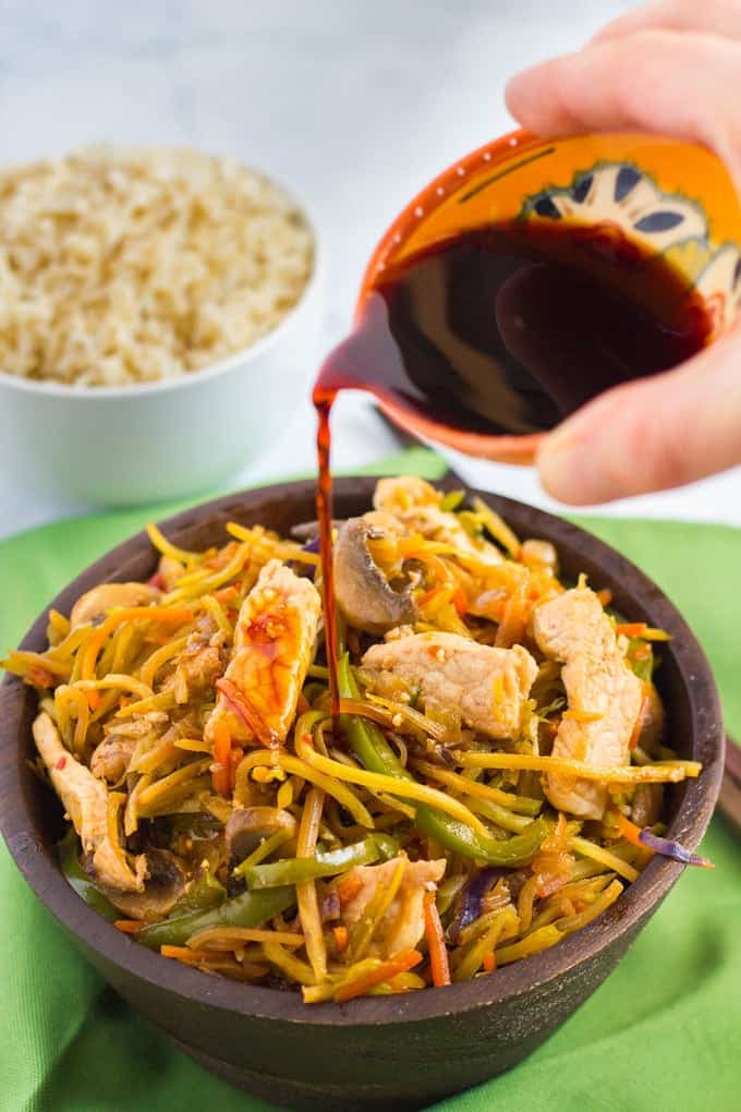 This veggie-packed pork stir fry is ready in just 25 minutes for a fast, healthy dinner! A hand pouring soy sauce from a small decorative bowl onto a pork stir fry mix in a wooden bowl.