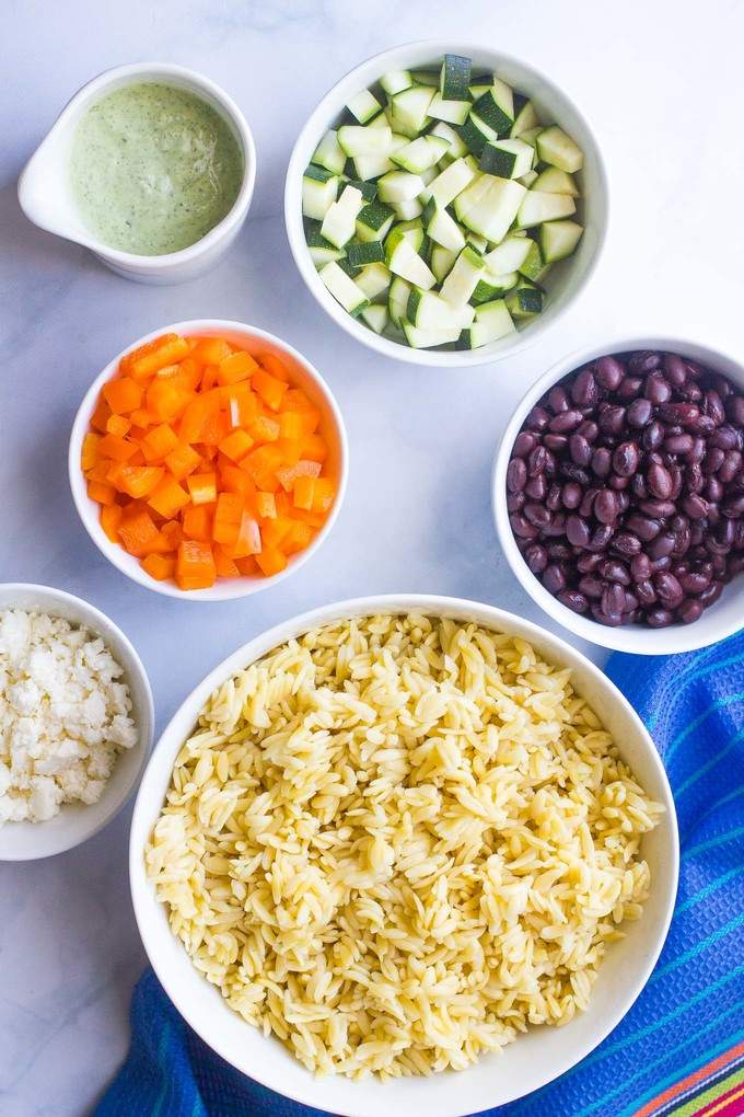 Separate bowls of orzo pasta, zucchini, black beans, chopped bell pepper, queso fresco and a creamy cilantro dressing