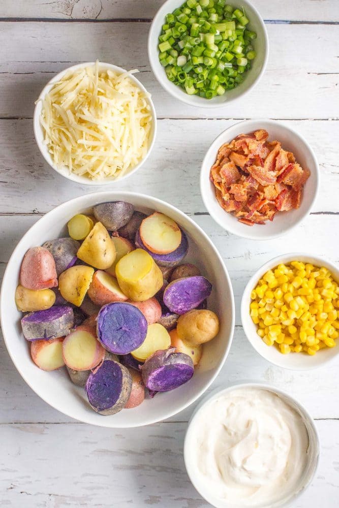 Ingredients laid out in separate bowls, including mixed color potatoes, cheddar cheese, bacon, corn, green onions and a creamy dressing