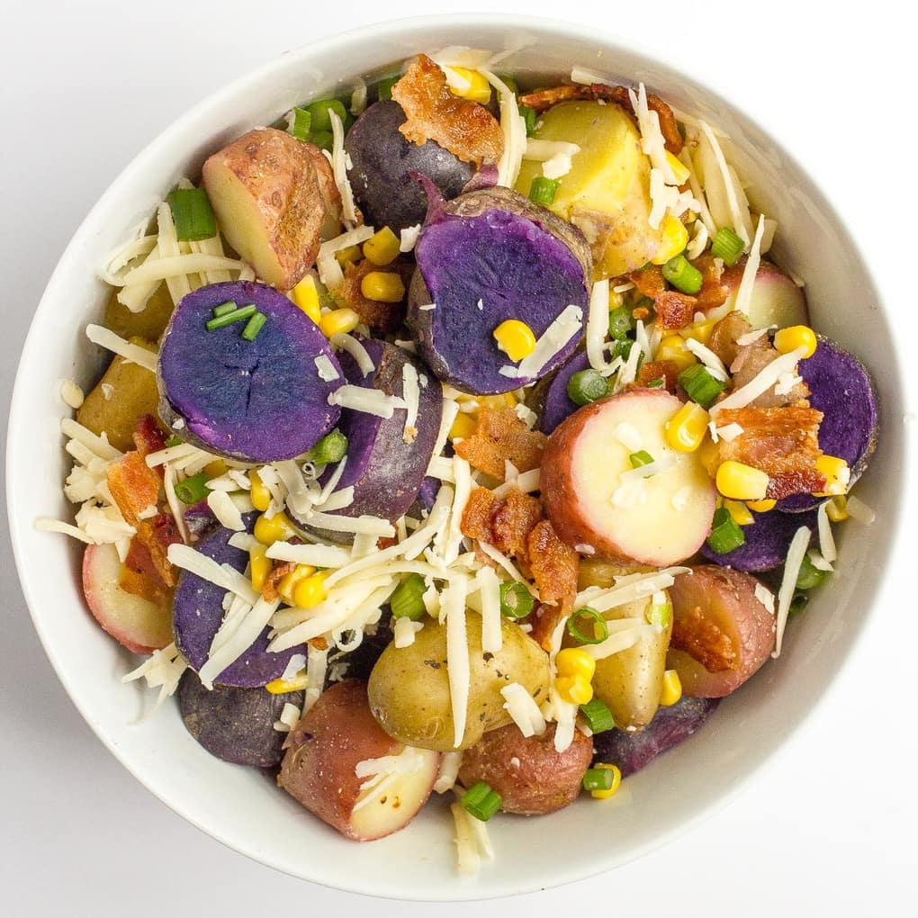 A white bowl with sliced red, yellow and purple potatoes along with cheddar cheese, corn and bacon