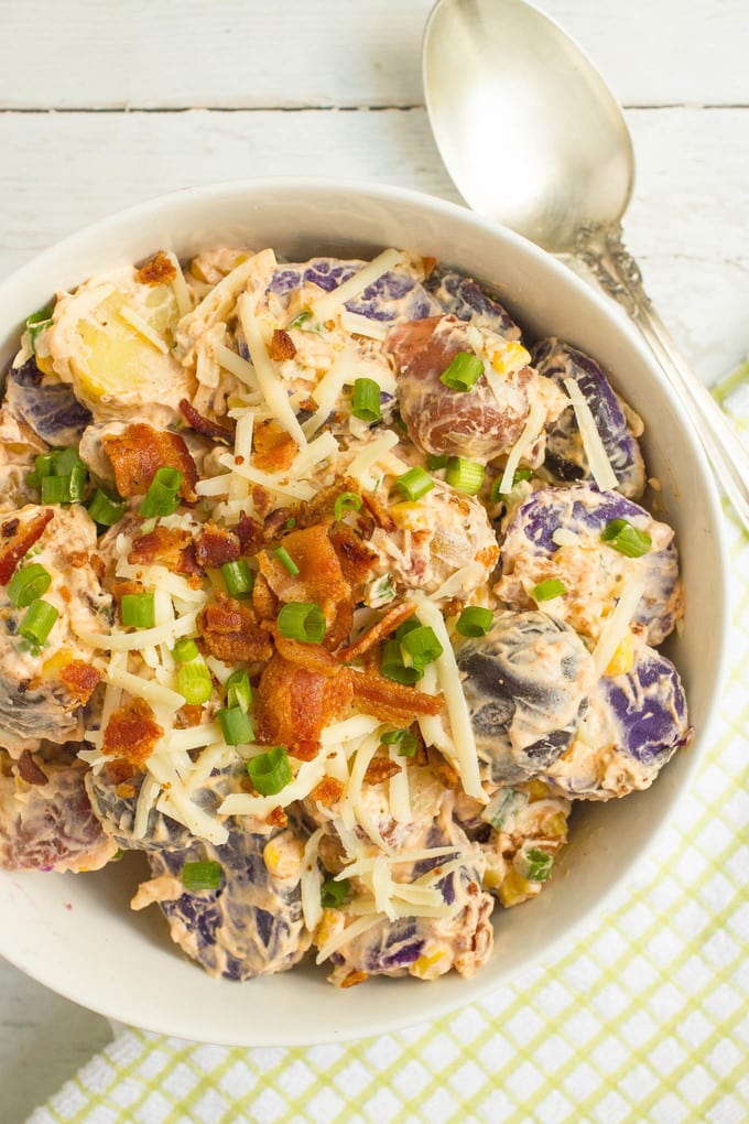 A creamy potato salad with cheddar cheese, corn and bacon mixed in, served in a large white bowl