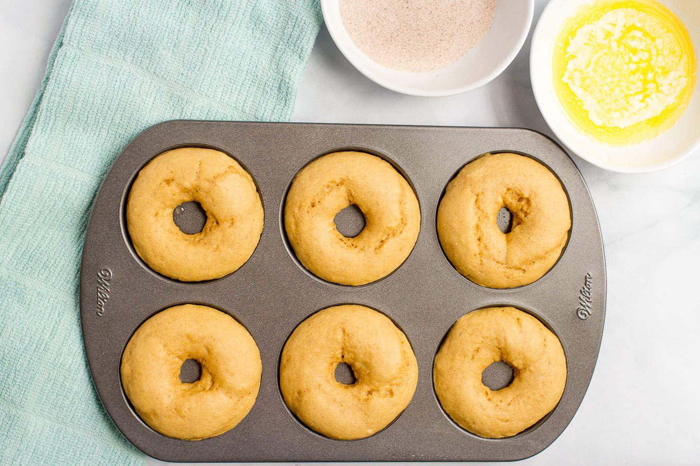 A nonstick donut pan with six baked donuts and bowls of butter and cinnamon sugar to the side.