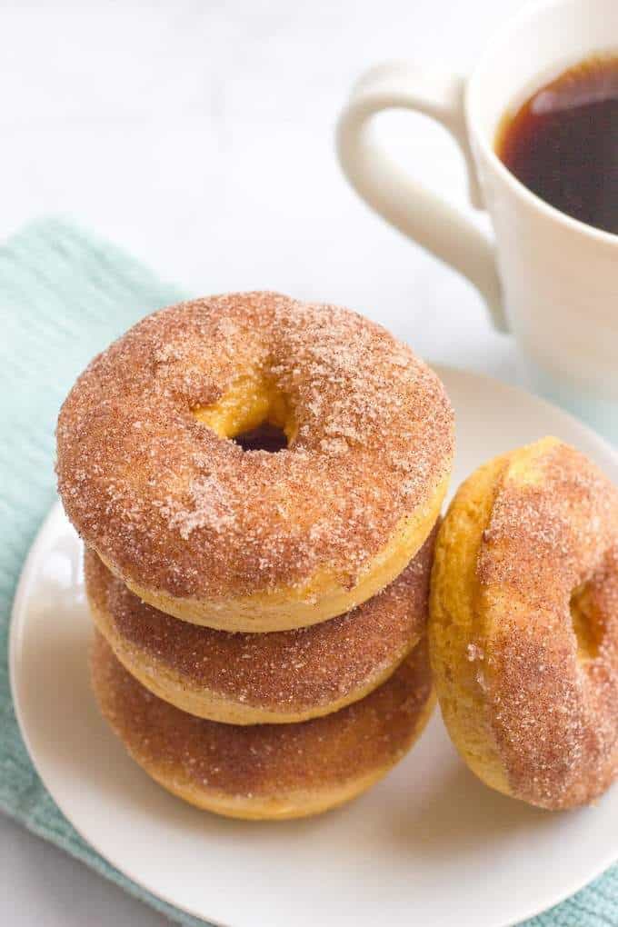 A stack of homemade baked cinnamon sugar donuts on a plate with a cup of coffee to the side.