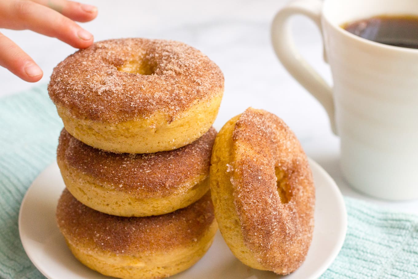 A small hand reaching for the top homemade baked donut on a stack of donuts on a white plate.