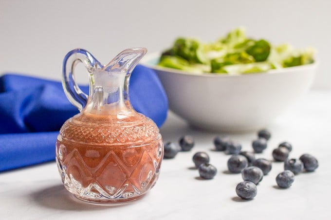 A small glass pitcher with a homemade blueberry vinaigrette with a salad bowl in the background and a smattering of blueberries nearby.