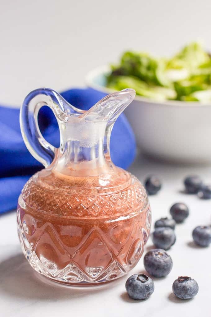 A small glass cruet with a blueberry vinaigrette with blueberries scattered to the side