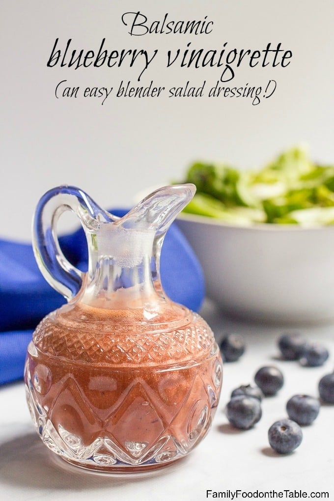 A glass cruet with balsamic blueberry vinaigrette with fresh blueberries to the side and a salad in the background with a text overlay on the photo