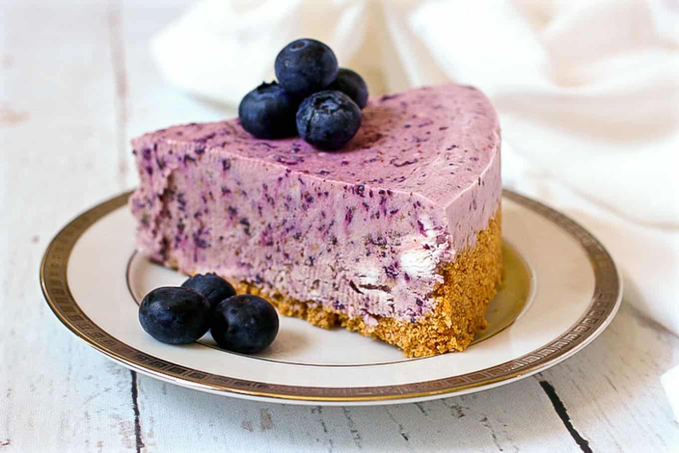 A tall, thick slice of a frozen blueberry pie with graham cracker crust on a china plate with fresh blueberries on top and to the side and a fork resting on the plate.