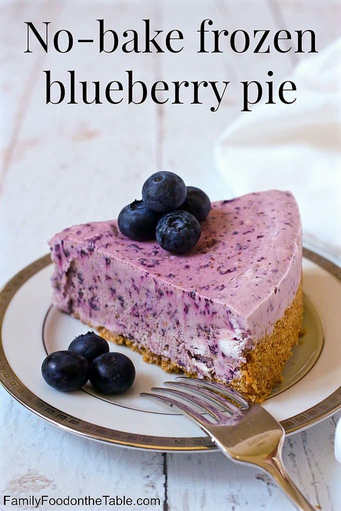 A slice of a frozen blueberry pie with graham cracker crust on a china plate with fresh blueberries on top and to the side and a fork resting on the plate.