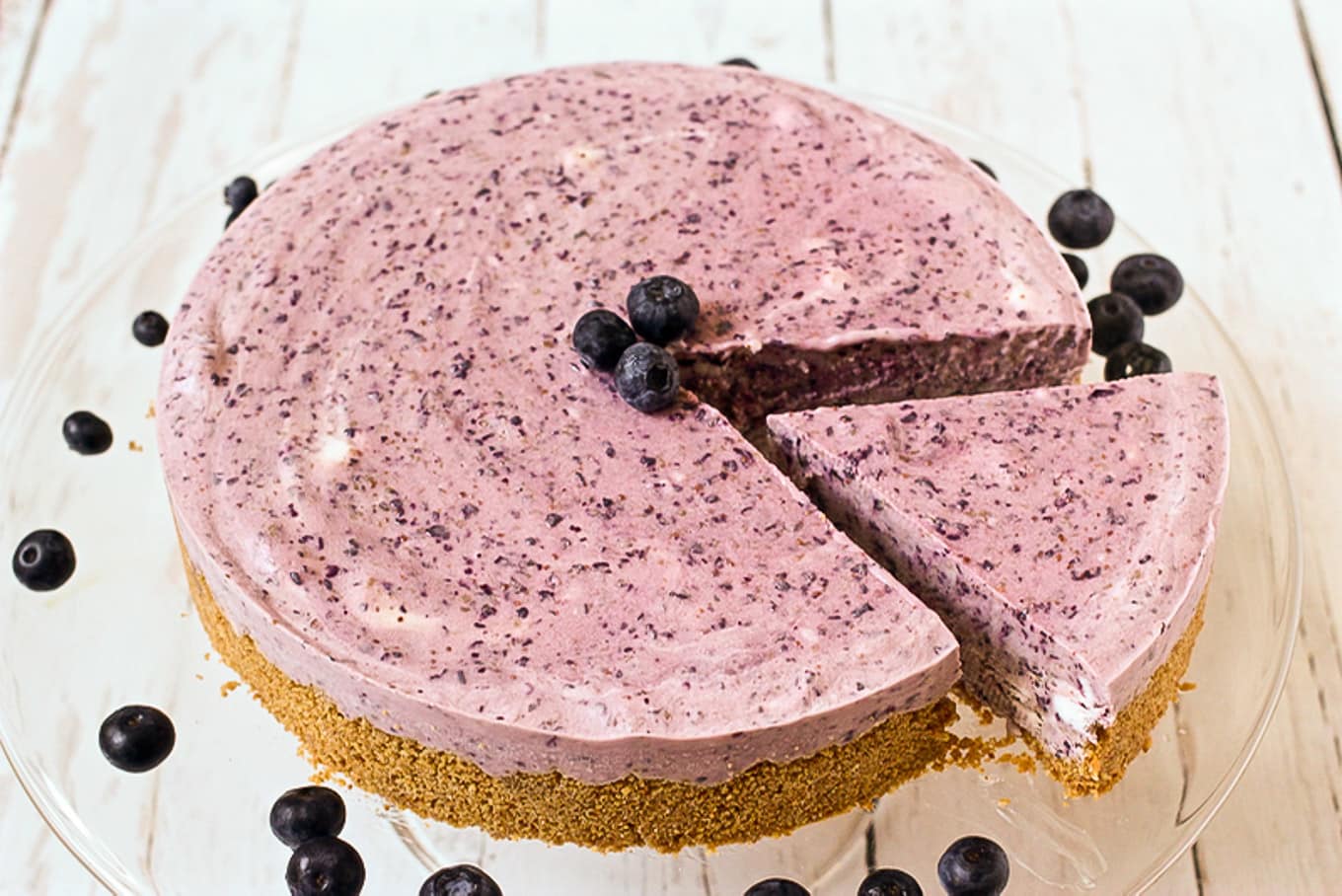 A slice of frozen blueberry pie being removed from the entire pie that\'s on a clear pie serving plate with fresh blueberries scattered about.