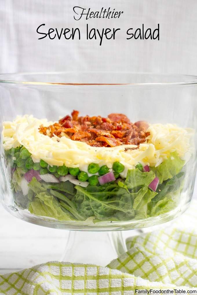 A clear tall glass trifle dish with a seven layer salad build in it and finished with bacon pieces on top.