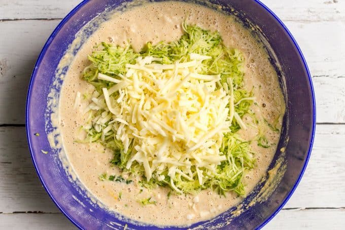 A large blue bowl with a waffle batter with shredded zucchini and shredded white cheddar cheese added on top before being mixed in.