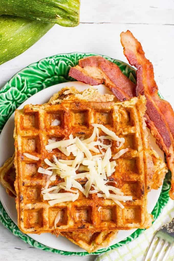 A plate with zucchini cheddar waffles topped with shredded white cheddar cheese with a couple of slices of bacon to the side.