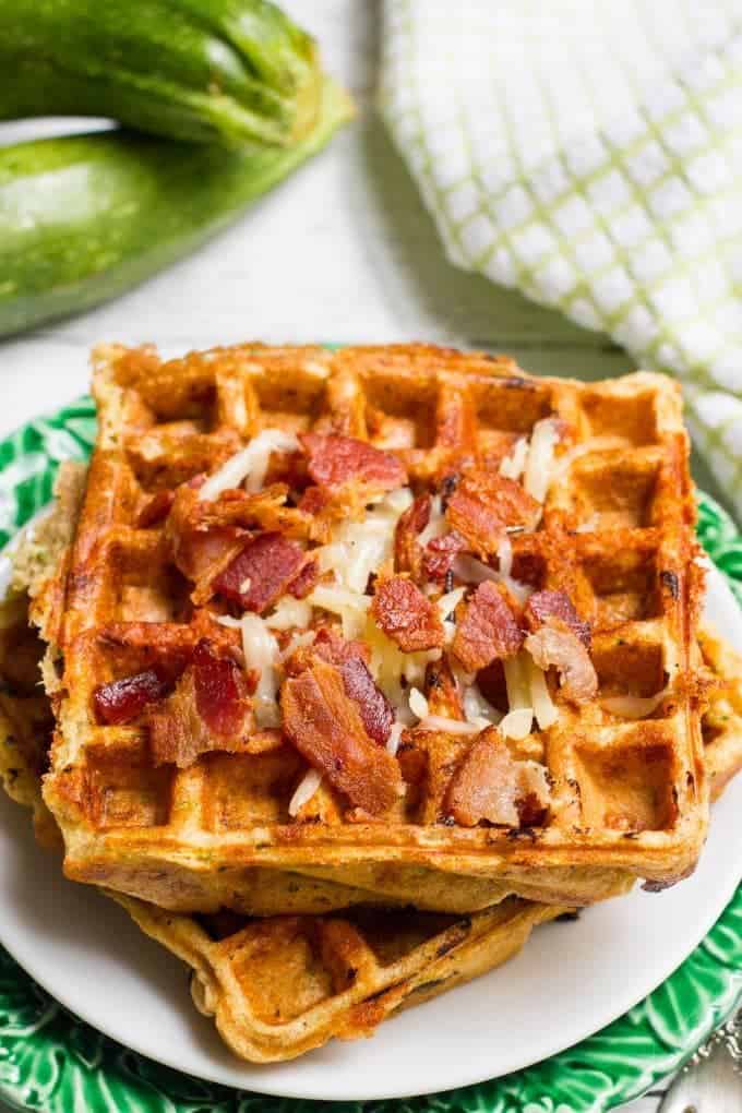 Zucchini waffles with cheese and bacon on top stacked and served on a plate with a zucchini in the background.