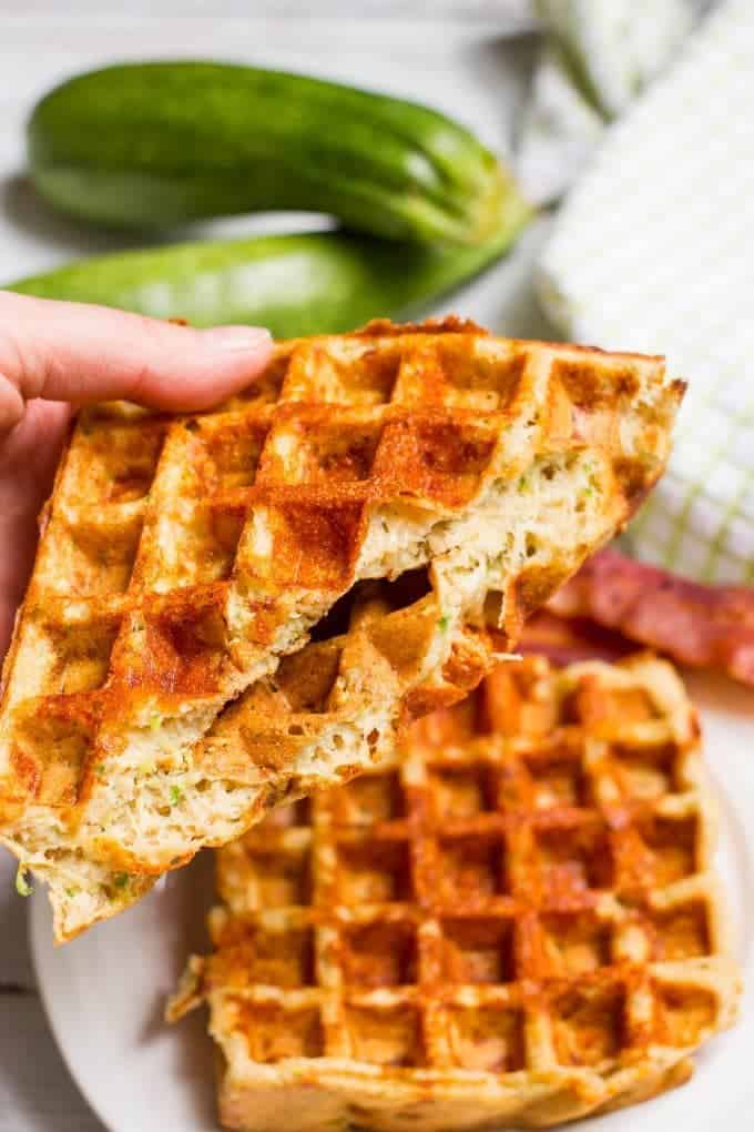A hand holding up a cheesy savory waffle folded in half from a plate.