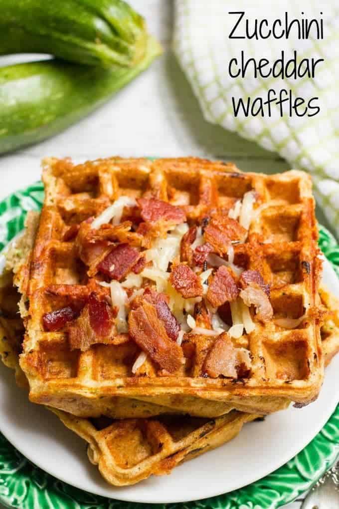 A plate with zucchini cheddar waffles topped with shredded white cheddar cheese and crumbled bacon.
