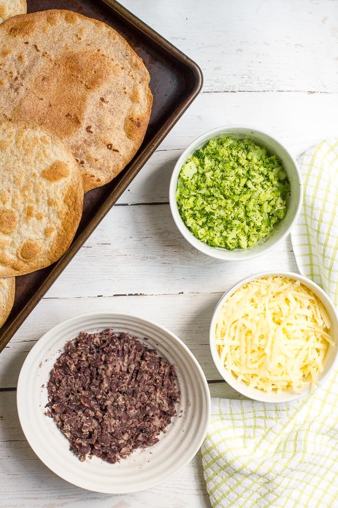 Toasted tortillas on a baking sheet alongside separate bowls of a mashed black bean mixture, finely chopped broccoli and shredded white cheddar cheese.