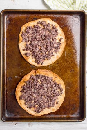 Two toasted tortillas on a baking sheet topped with a partially mashed black bean mixture/