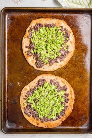 Two tostadas on a baking sheet topped with mashed black beans and finely chopped broccoli.