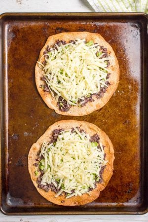 Two tostadas on a baking sheet topped with mashed black beans and finely chopped broccoli and shredded white cheddar cheese before being melted.