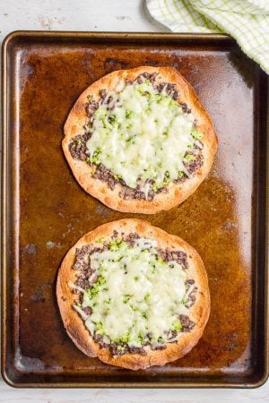 Two tostadas on a baking sheet topped with mashed black beans and finely chopped broccoli and melted shredded white cheddar cheese.