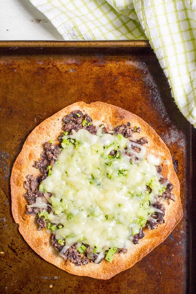 Close up of a black bean tostada with chopped broccoli and melted white cheddar cheese on a baking sheet.